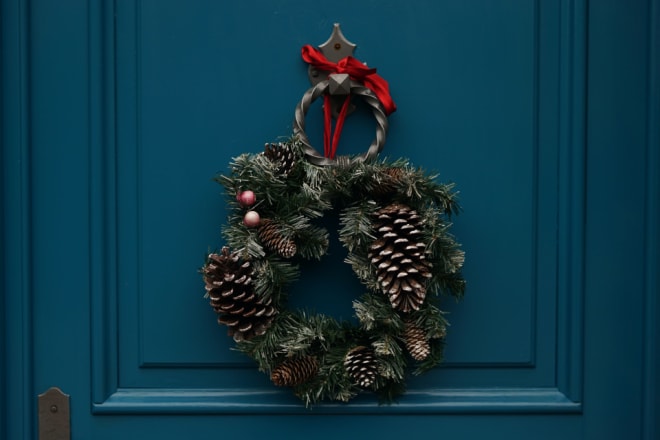 A holiday wreath made of evergreens and pinecones hangs from a round metal doorknocker attached to a teal door.